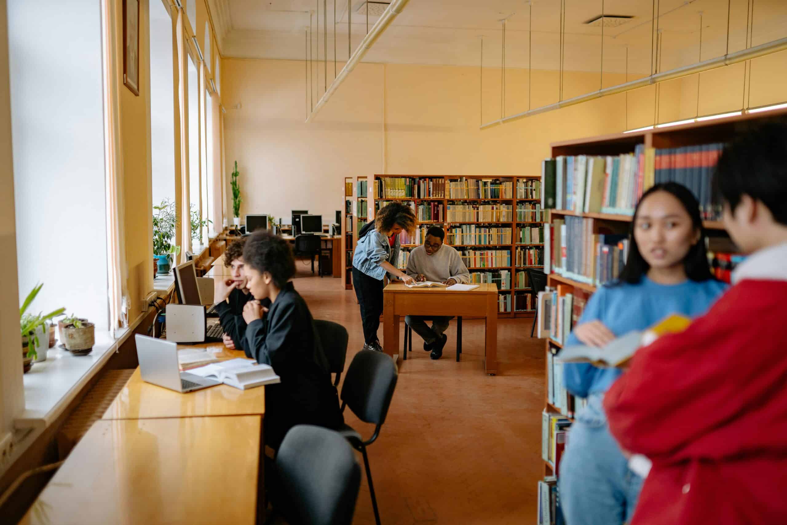 Students studying and reading in a well-lit library with bookshelves and computers.